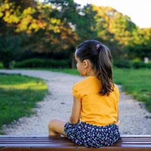 Una bambina di circa 8 anni è seduta di spalle su una panchina di legno in un parco